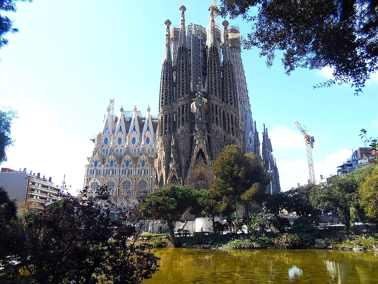 The basilica and its tall towers rising behind the trees and the pond of the small park, with a crane at the right