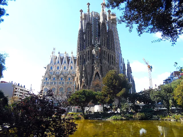 The basilica and its tall towers rising behind the trees and the pond of the small park, with a crane at the right